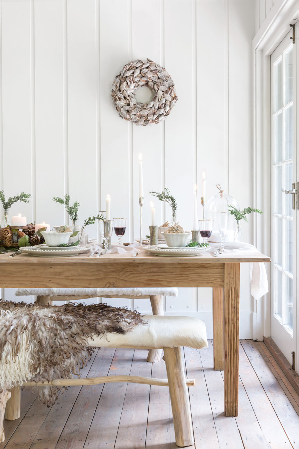 A wooden dining table set for a meal with white plates, glasses, candles, and greenery showcases elegant interior design. A rustic bench with a textured throw sits in front, whilst a wreath adds charm to this Dorset-inspired residential styling.