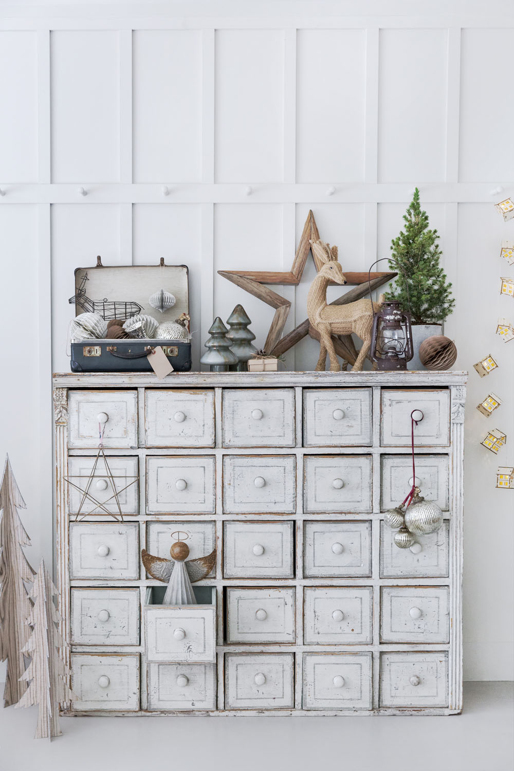A distressed white cupboard with many drawers is decorated with Christmas baubles, small trees, a star, a lantern, and a wooden reindeer. The festive scene evokes the cosy charm of Dorset, set against white panelled walls and gold fairy lights.