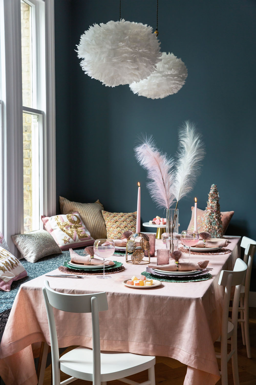 A dining table with a pale pink tablecloth is set for a meal with pink and green plates, pink glasses, candles, and pampas grass centrepieces. White chairs surround the table; cushions line a window seat against a dark blue wall.