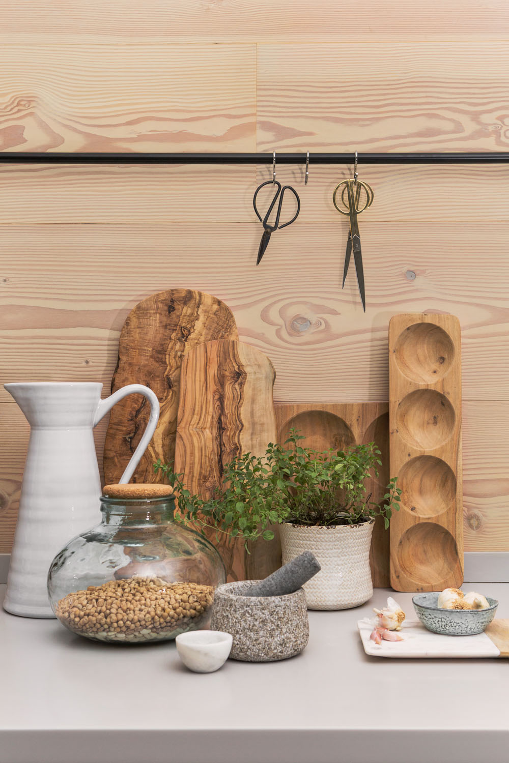 A kitchen worktop with a white jug, a glass jar filled with chickpeas, a stone mortar and pestle, sliced ginger, small bowls, a potted plant, wooden boards, and two hanging scissors against a wood-panelled wall.