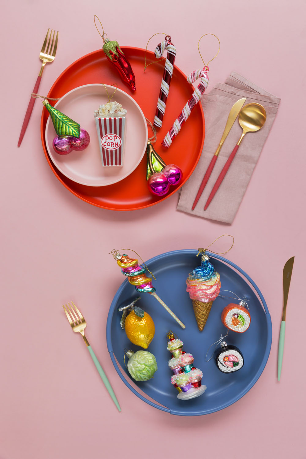 Two plates on a pink surface, one red with cutlery and glass ornaments shaped like popcorn, chilli, and fruit; one blue with cutlery and ornaments shaped like sushi, ice cream, pizza, and sweets.