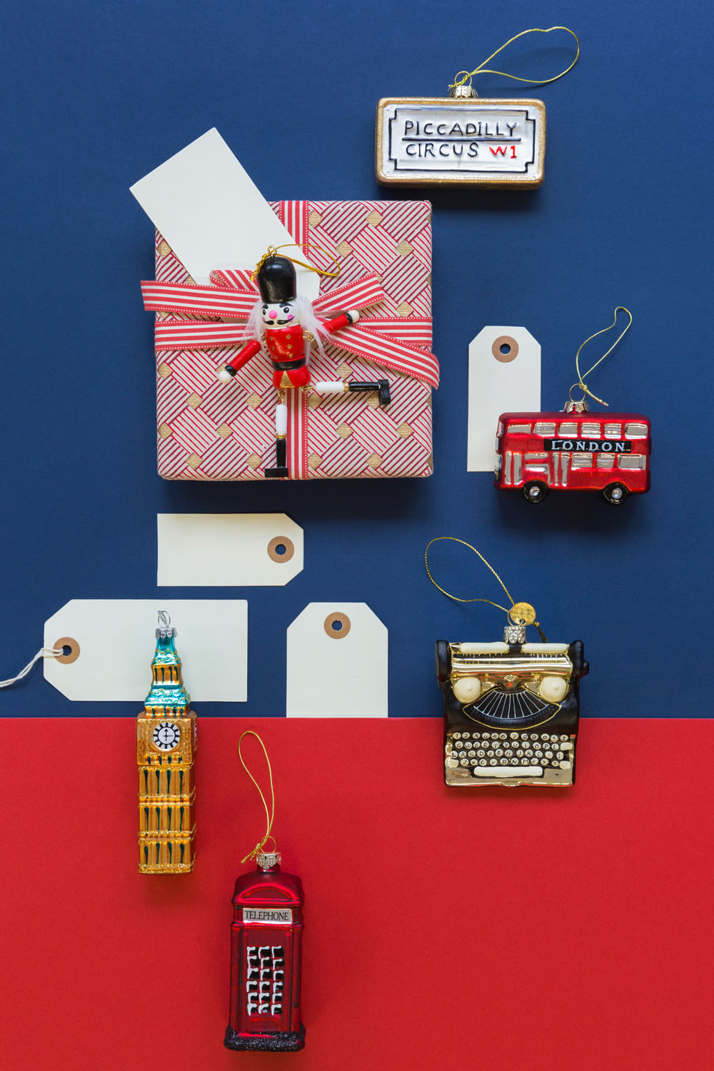 A wrapped present with a toy British guard on top sits among blank tags and London-themed ornaments: a double-decker bus, a Piccadilly Circus sign, Big Ben, a typewriter, and a red telephone box, on a red and blue background.