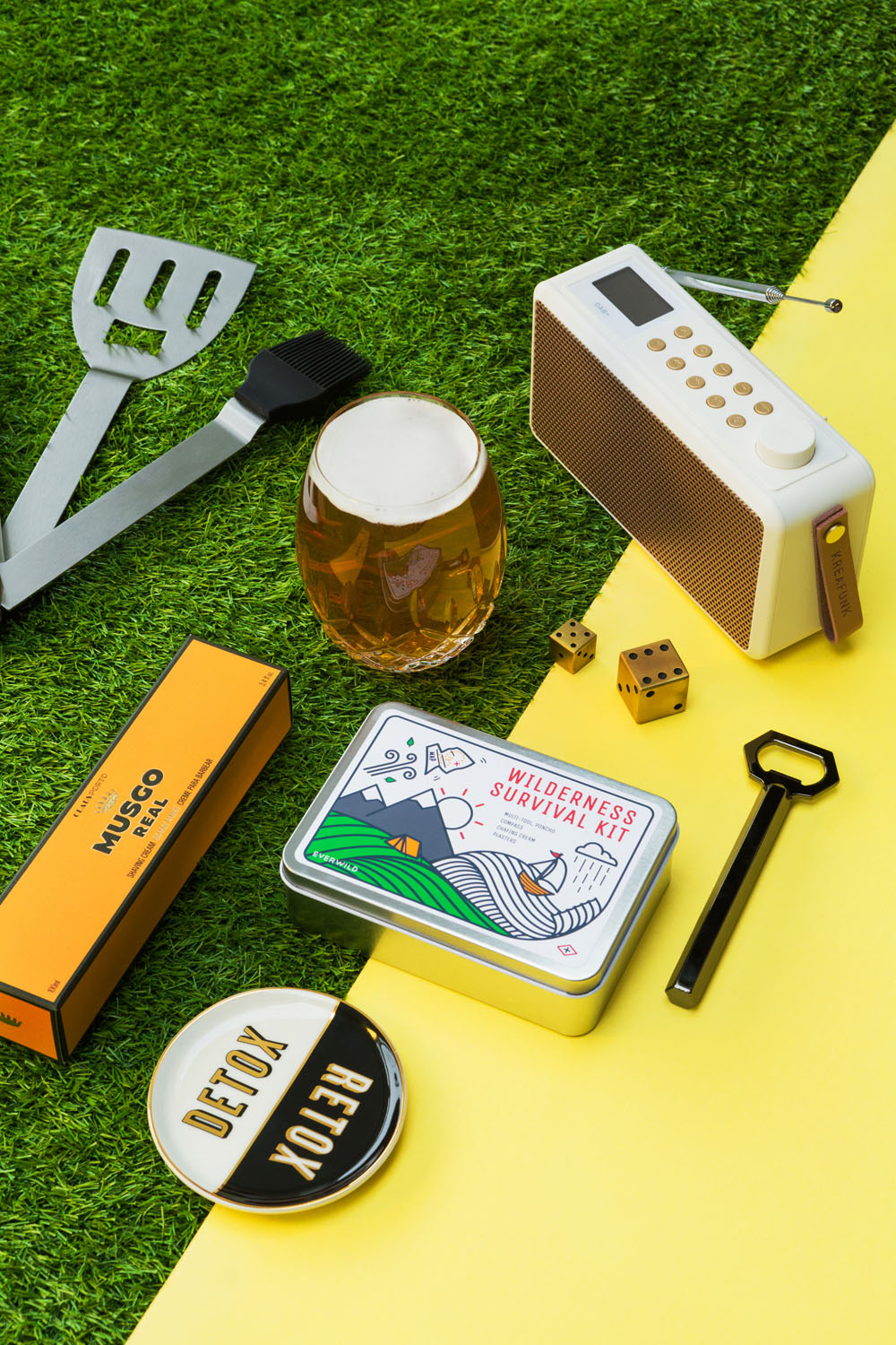 A flat lay on a green and yellow background displays a spatula, grill brush, radio, glass of beer, dice, bottle opener, tin labelled Wilderness Survival Kit, small boxes, and round tins labelled MUSCO, RETRO, and COED.