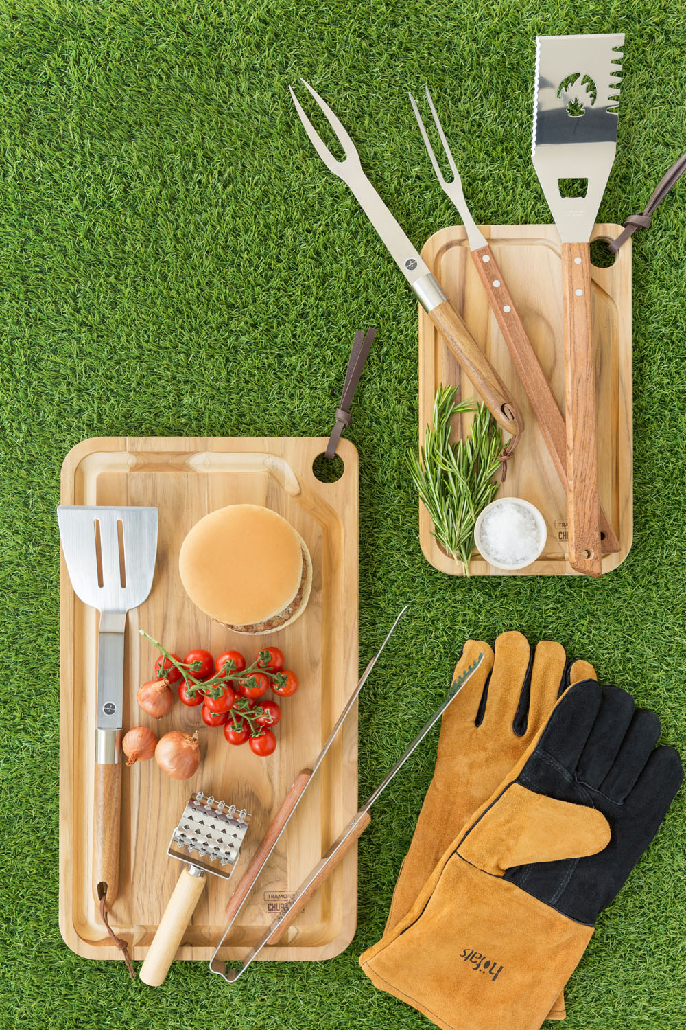 A top-down view of grilling tools, a pair of brown gloves, a chopping board with a burger bun, cherry tomatoes, onions, and herbs, all arranged on green artificial grass.