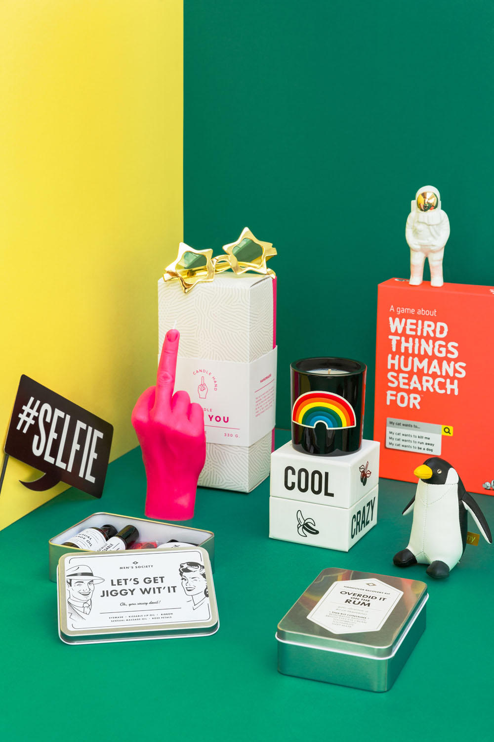 A colourful arrangement of novelty gifts on a split yellow and green background, including a pink hand sculpture showing the middle finger, a rainbow mug, a penguin figure, tins, an astronaut figure, and a #SELFIE sign.