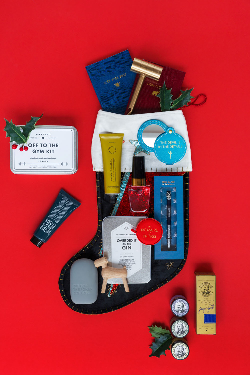 A black Christmas stocking on a red background is filled with various self-care and gift items, including skincare products, a red nail varnish, hand creams, and other small gifts. Holly sprigs lie beside the stocking.