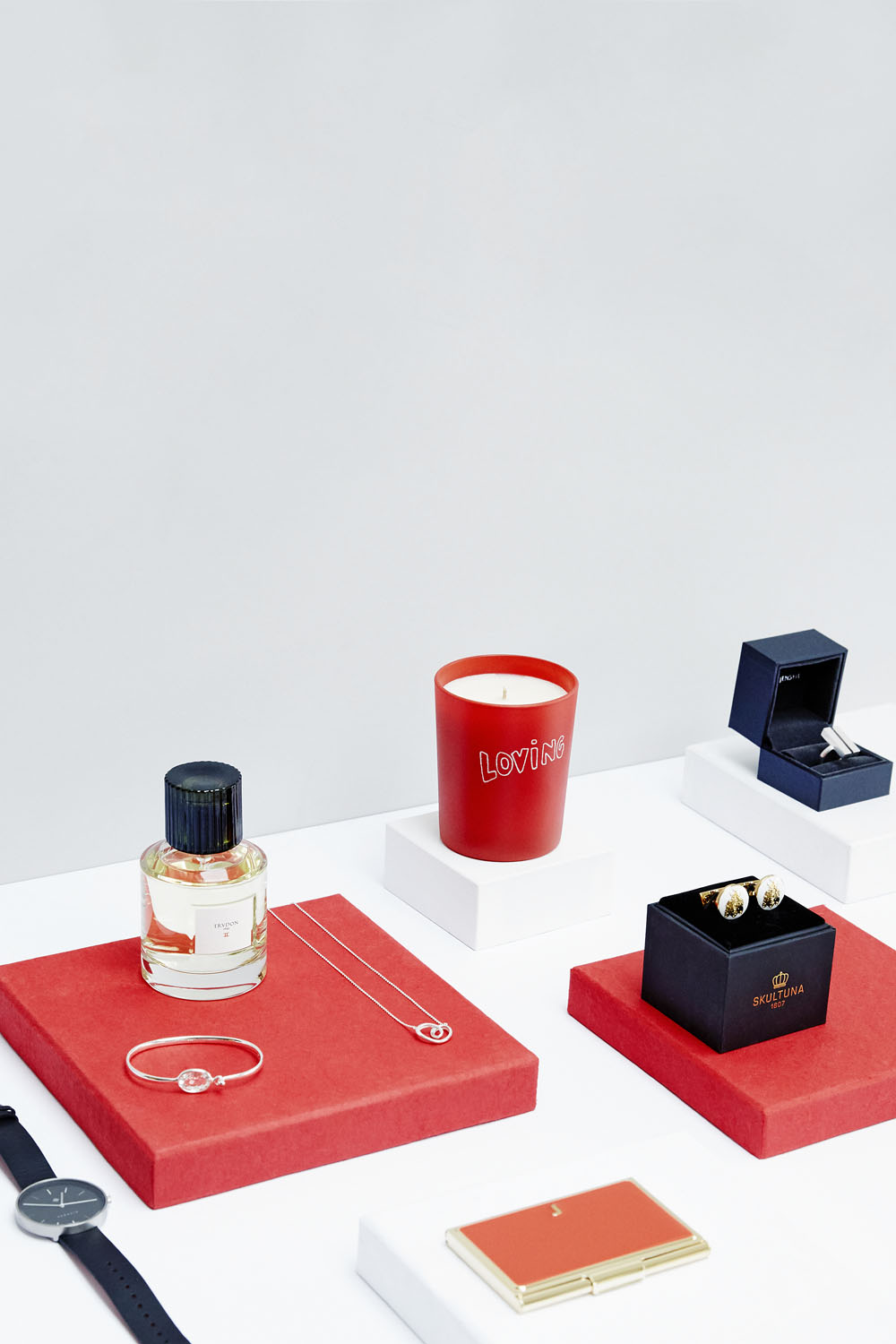 A collection of luxury items displayed on red and white plinths, including a red candle, a perfume bottle, a watch, cufflinks in a box, a necklace, a bracelet, and a gold card holder.