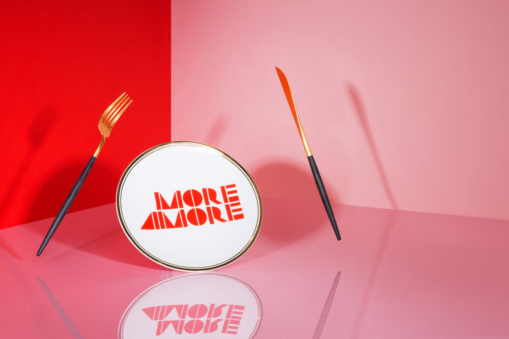 A plate with MORE AMORE in bold red letters sits on a reflective pink surface, flanked by a gold and black fork and knife. The background features red and pink colour blocks.