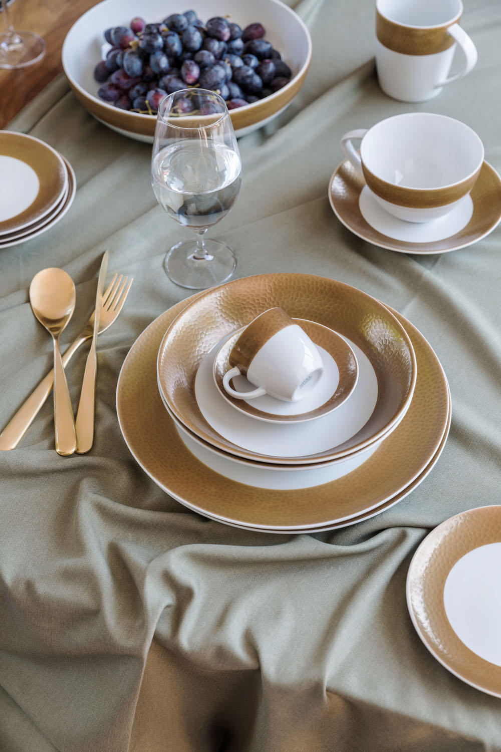 A table set with gold and white crockery, including plates, cups, and saucers, gold cutlery, a glass of water, and a bowl of grapes, arranged on a green tablecloth with gentle folds.