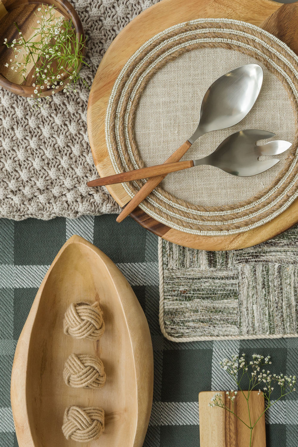 A tabletop arrangement with a wooden plate, placemat, salad servers, wooden tray holding three rope knots, coasters, a bowl of salt, and sprigs of baby's breath on a chequered green tablecloth.
