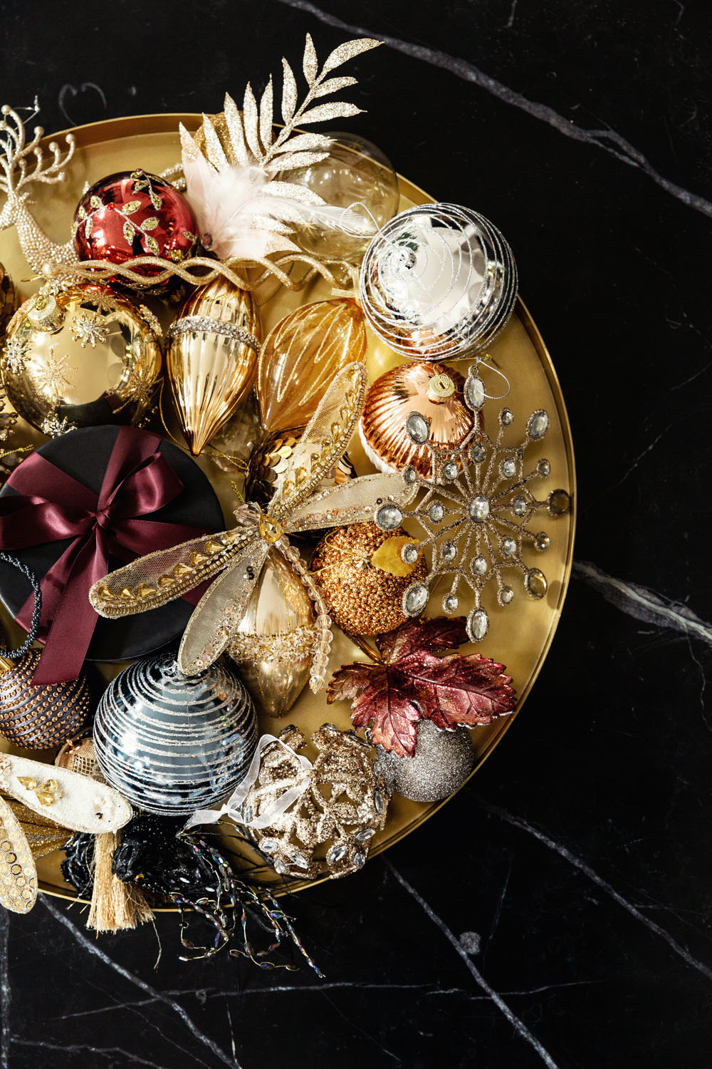 A gold tray on a black marble surface holds assorted Christmas baubles, including gold, silver, red, and glittery decorations, a snowflake, a dragonfly, leaves, and a black box with a burgundy ribbon.
