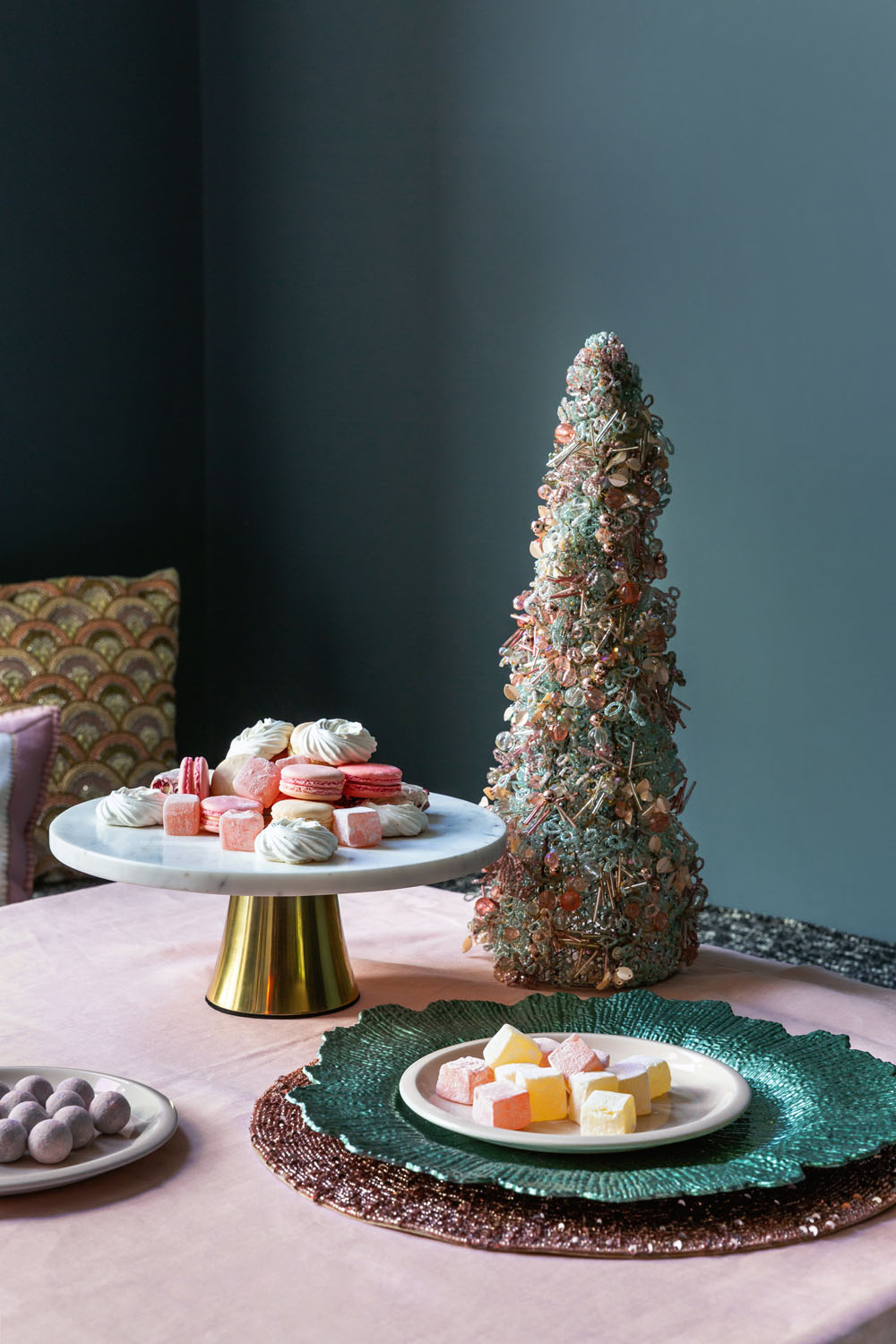 A table displays assorted pastel-coloured sweets on plates and a cake stand, next to a tall, decorative cone-shaped centrepiece resembling a Christmas tree, with a dark teal wall and patterned cushions in the background.