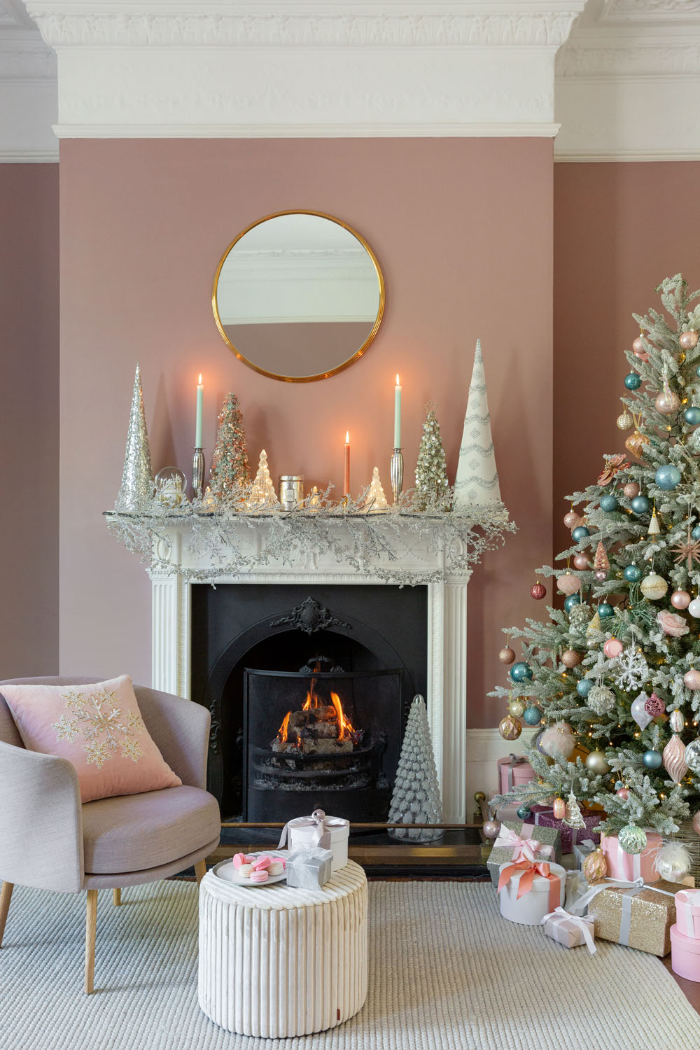 A living room with a lit fireplace, decorated mantelpiece with candles and cone ornaments, a round mirror above, a Christmas tree with pastel baubles, wrapped presents underneath, a pink cushion chair, and a round ottoman with a tray.