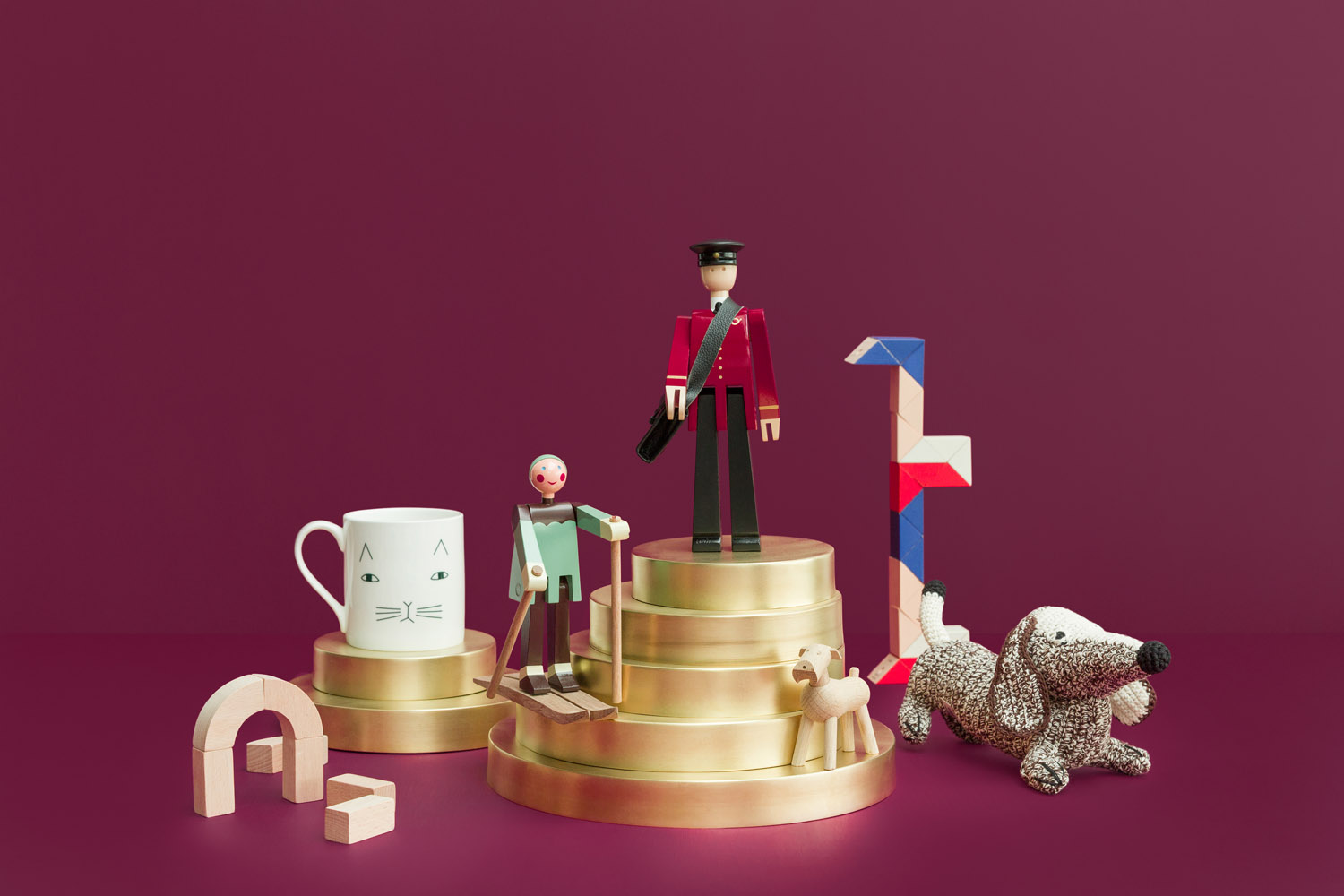 A group of objects on gold cylindrical podiums against a burgundy background, including a white mug with a cat face, wooden human figurines, a toy dog, wooden building blocks, and a small arch.