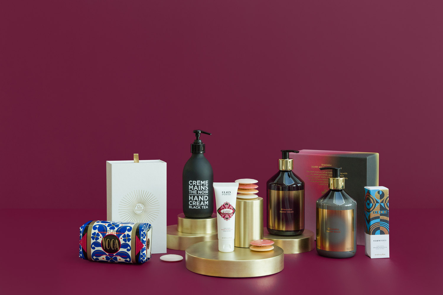 A variety of skincare and beauty products, including bottles, tubes, and boxes, are arranged on round gold platforms against a maroon background. The items appear neatly organised for a display or advertisement.