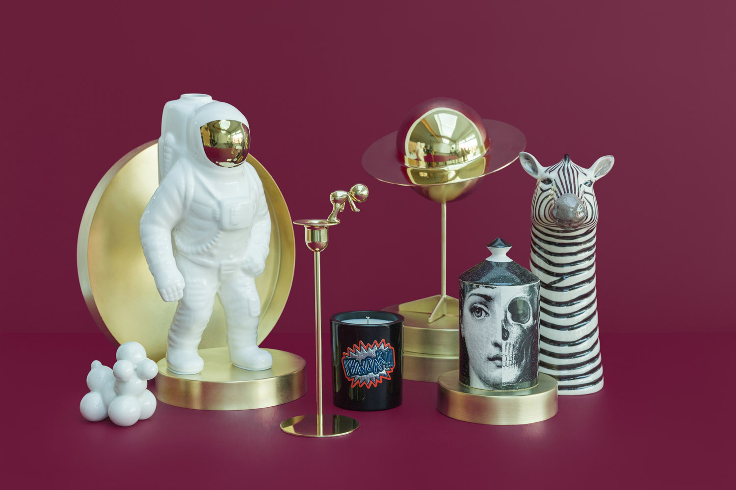A group of decorative objects on gold platforms against a burgundy background includes a ceramic astronaut, a gold planet sculpture, a zebra figurine, a face-printed container, a candle, and a cluster of white spheres.