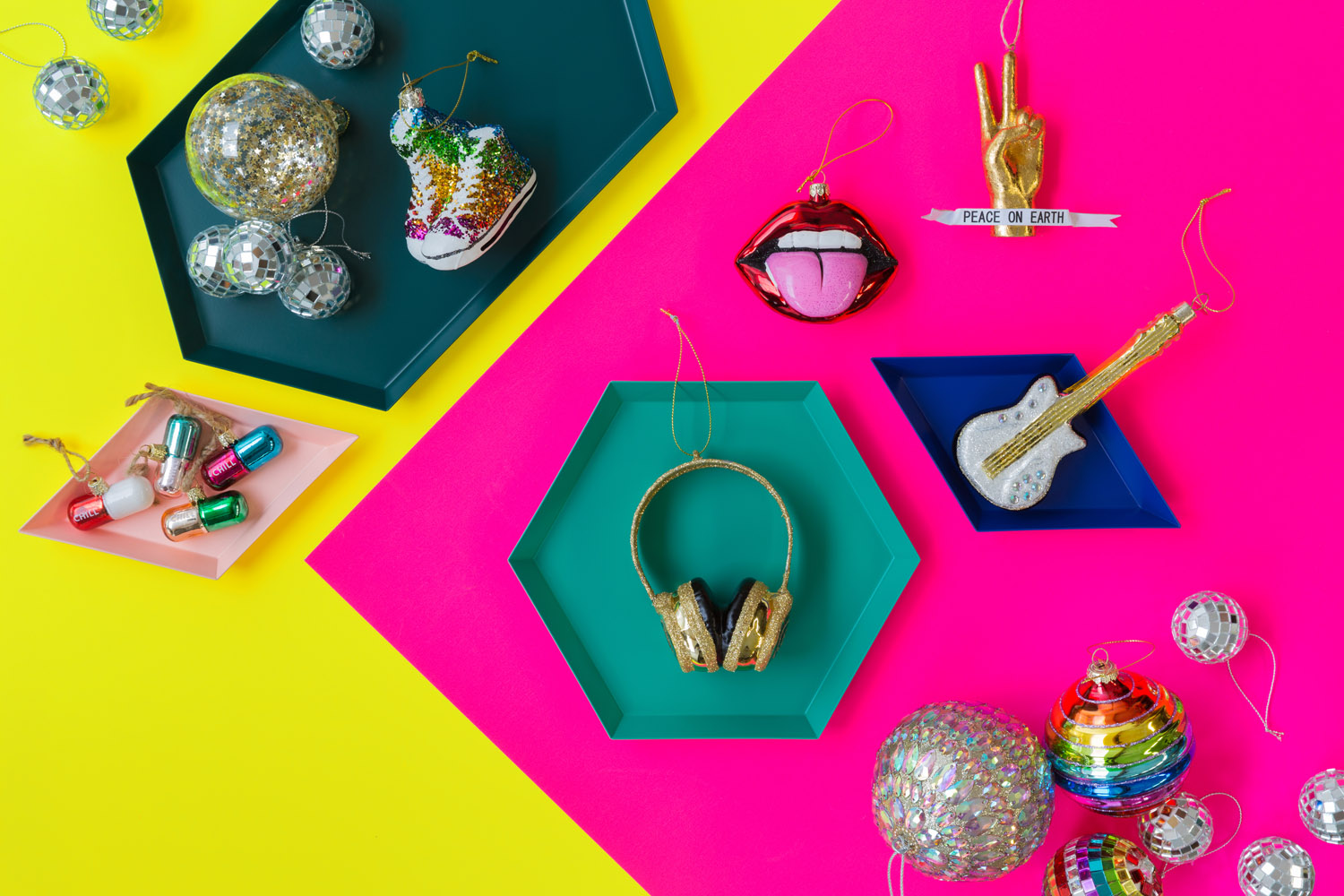 Colourful flat lay of various Christmas ornaments on yellow and pink backgrounds, including disco balls, pills, a headphone, guitar, lips with tongue out, a rainbow ball, a hand peace sign, and a pine tree.
