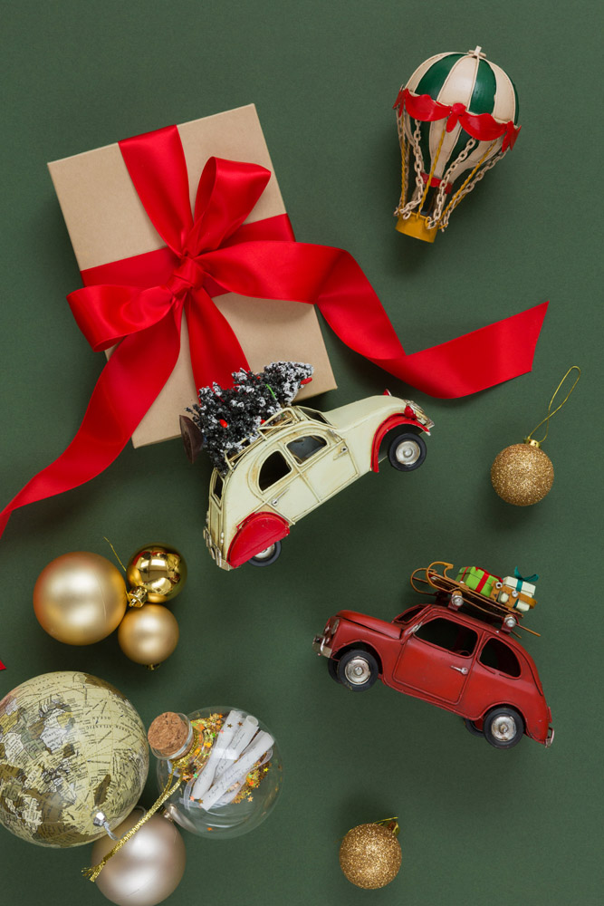 Gift box with red ribbon, two toy cars carrying Christmas trees and presents, gold and glitter ornaments, a small decorative hot air balloon, and a glass jar on a green background.
