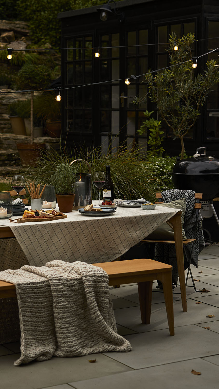 A wooden outdoor table with a checked tablecloth is set with plates, glasses, wine, snacks, and breadsticks. Benches, a knitted throw, fairy lights, plants, and a black barbecue are visible in a garden setting.