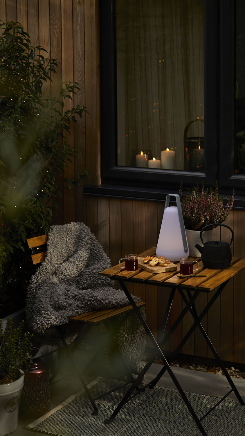 A small wooden table and chairs set on a patio with a cosy grey blanket, tea, biscuits, a black teapot, a glowing lantern, and potted plants, with candles visible inside a nearby window.