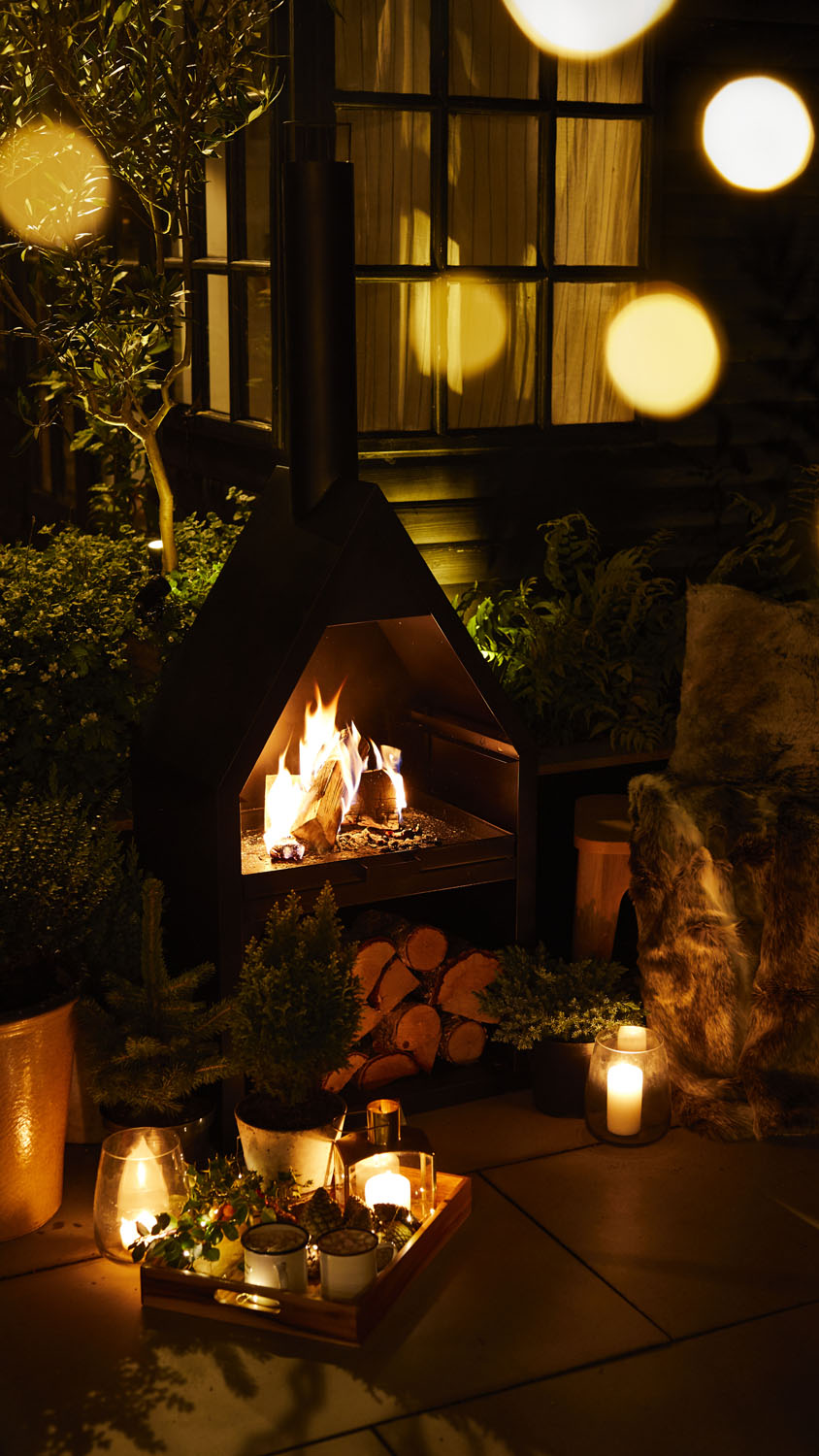 An outdoor fireplace with a burning fire is surrounded by logs, plants, candles, and a tray with drinks and snacks, set on a patio at night with soft lighting and blurred lights in the foreground.