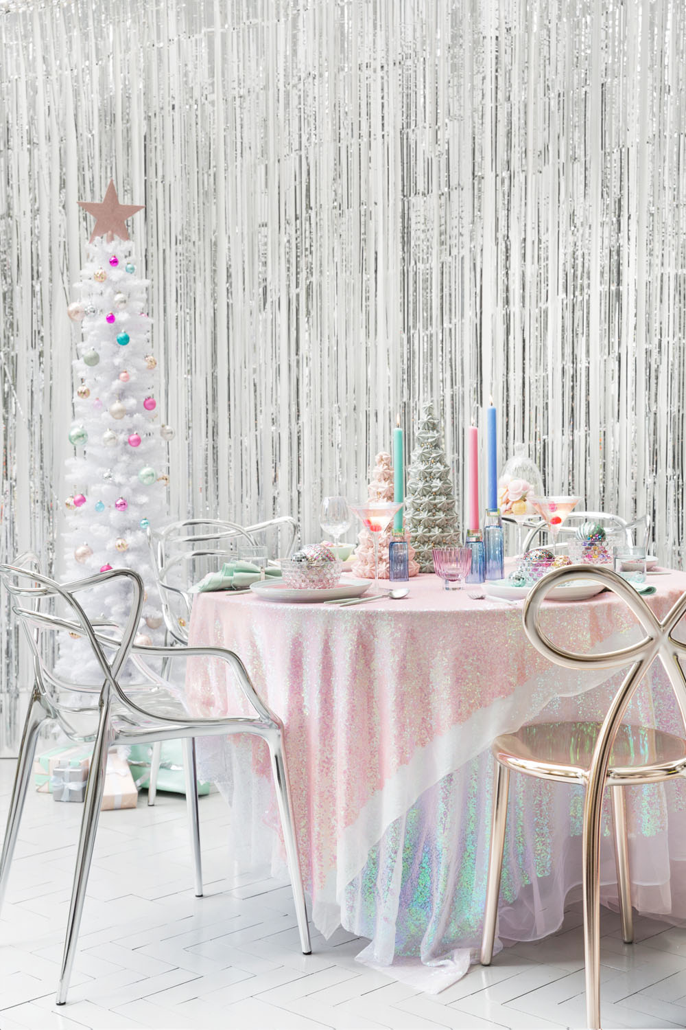 A round table with a shimmery pink iridescent tablecloth is set with pastel candles, glassware, and holiday decorations. Transparent chairs surround the table. A white Christmas tree is in the background with a silver tinsel backdrop.