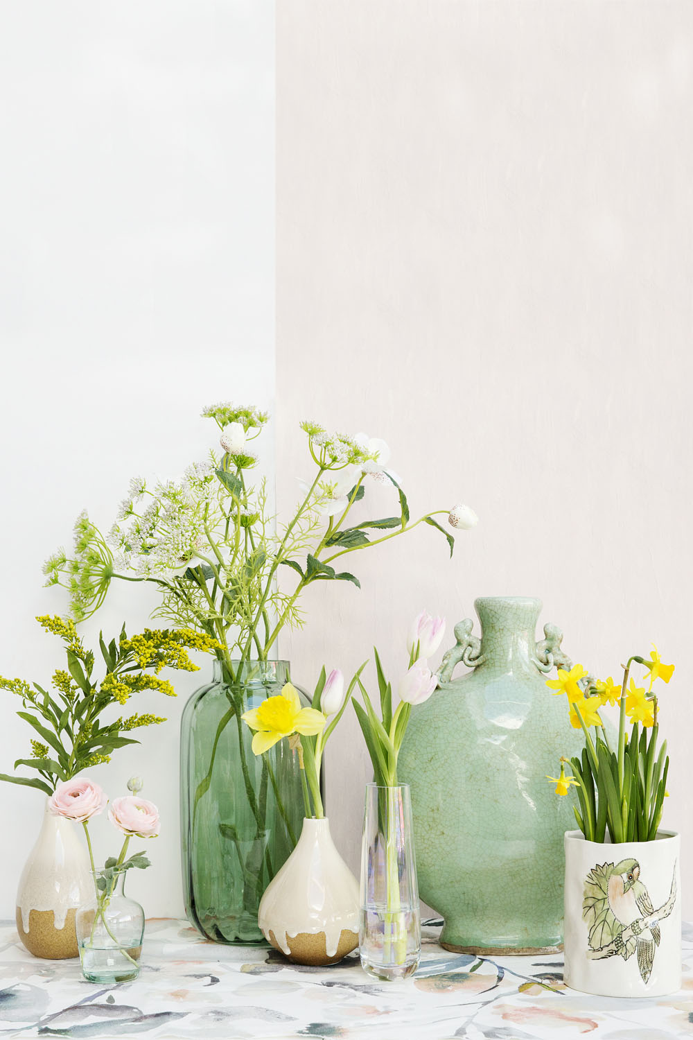 Several vases and vessels, some green and some off-white, display a variety of fresh spring flowers, including tulips, daffodils, and daisies. The arrangement sits on a marble-patterned surface against a pale wall.