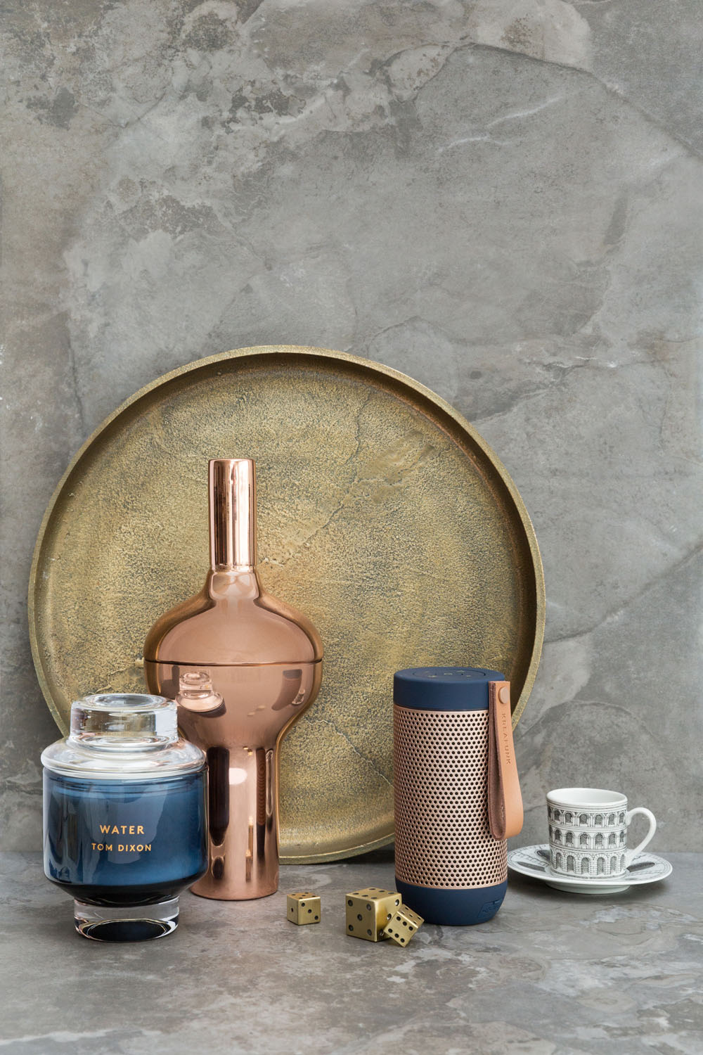 A gold tray, a copper vase, a blue candle labelled Water, two gold dice, a portable speaker with a leather strap, and a teacup with a saucer are arranged on a grey stone surface.