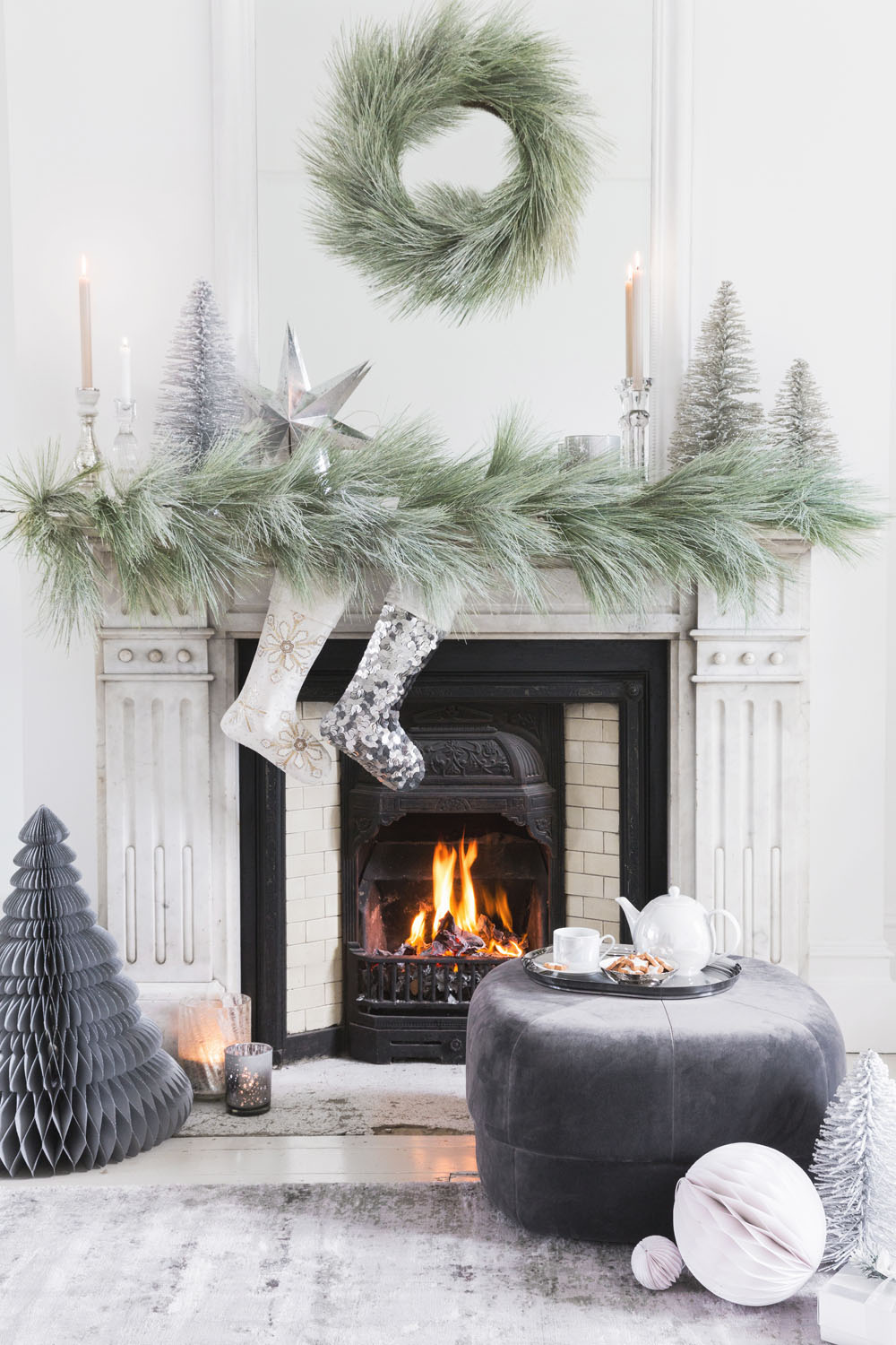A decorated white fireplace with a green garland, silver trees, two stockings, and a wreath. A fire burns in the fireplace. Nearby are a grey ottoman, tea set, paper trees, and a white ornament on a light rug.