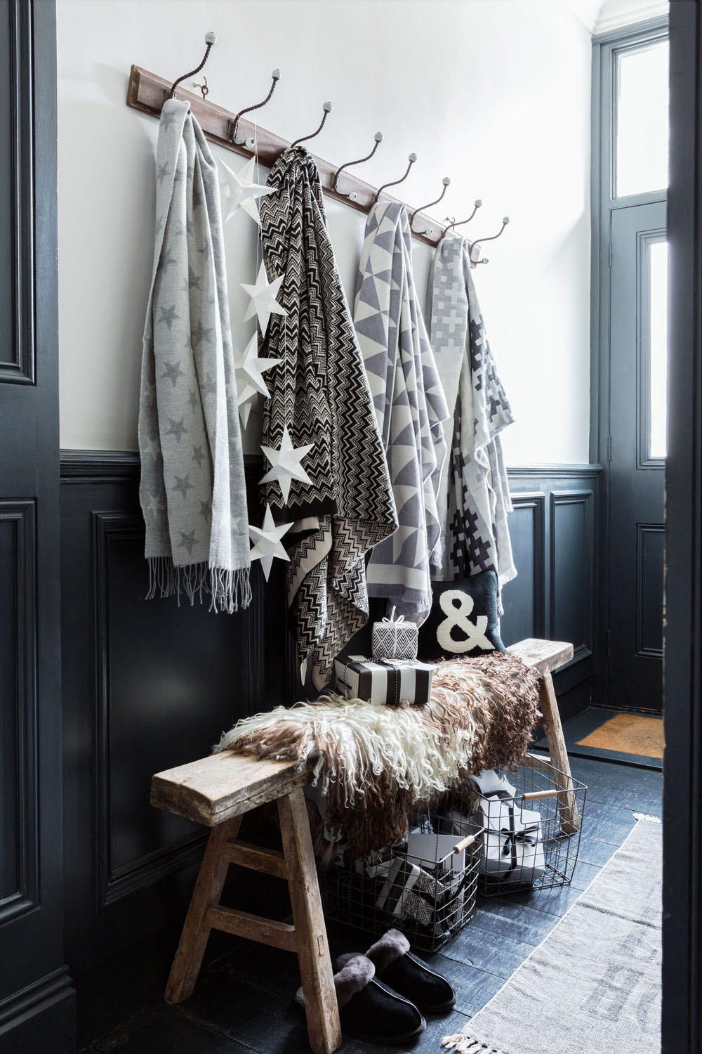 A wooden bench sits below a coat rack with six patterned grey and white blankets. Wrapped presents and a wire basket are on the bench and floor. Decorative accents and slippers are also visible in the entrance hall.