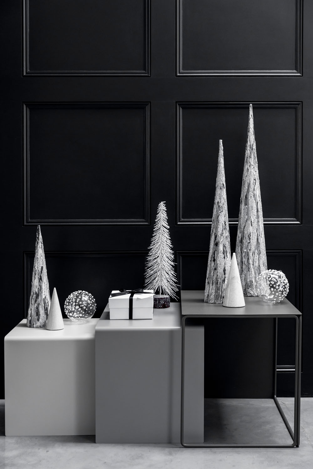 Three modern nesting tables display cone-shaped decorative trees, two decorative spheres, and a wrapped gift box with a black ribbon, set against a black panelled wall in a minimalist, monochrome style.