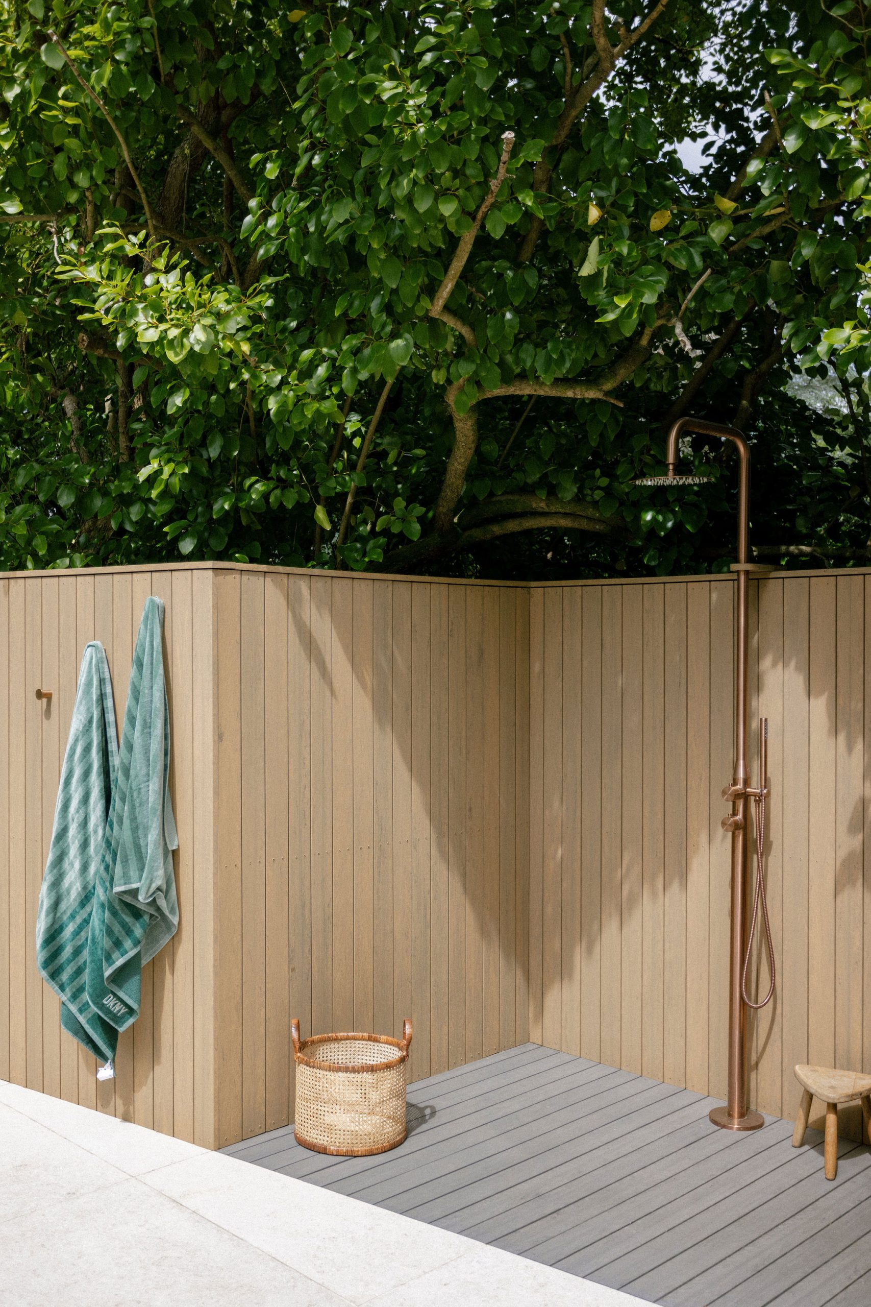 An outdoor shower area with wooden panelled walls, two green towels hanging on hooks, a woven basket on the ground, and a leafy tree overhead. A bronze shower fitting and a small wooden stool are also present.