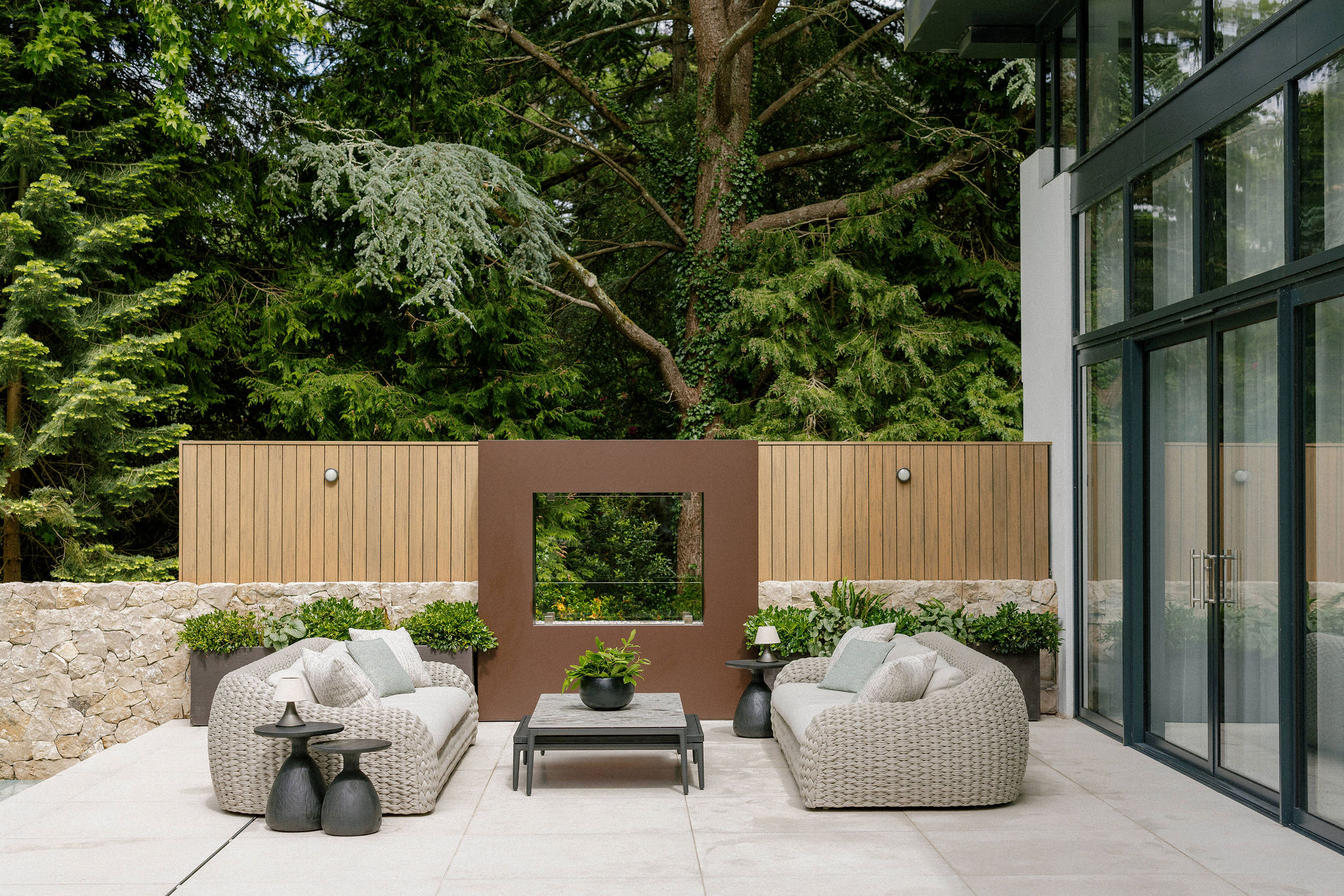 Modern outdoor patio with two woven sofas facing each other, a coffee table in the centre, potted plants, and stone and wood privacy fencing. Large glass doors lead inside, with tall trees in the background.