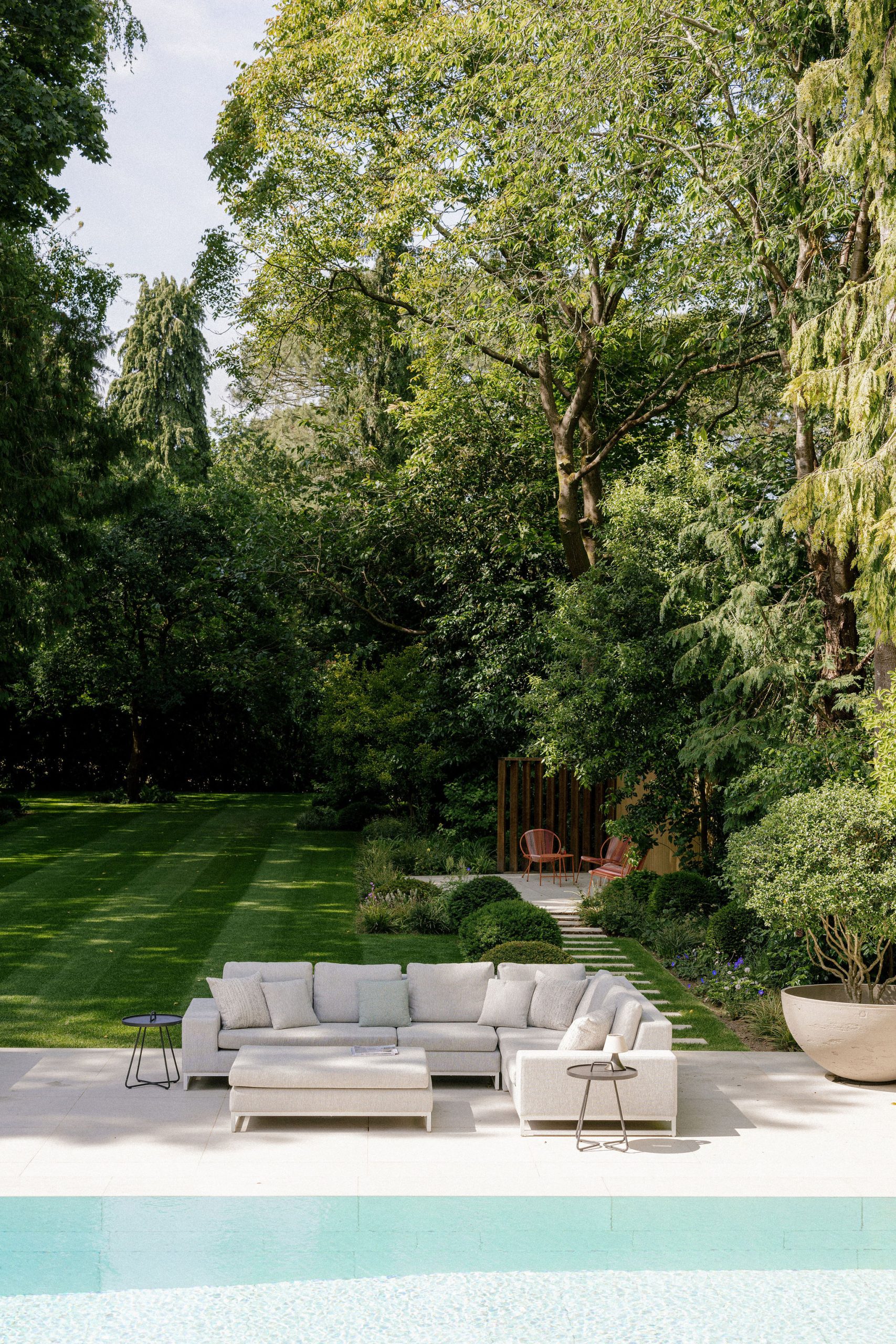 A modern outdoor seating area with light grey sofas and side tables is set on a patio by a swimming pool. Lush green grass, trees, and a small garden seating area with two pink chairs are visible in the background.