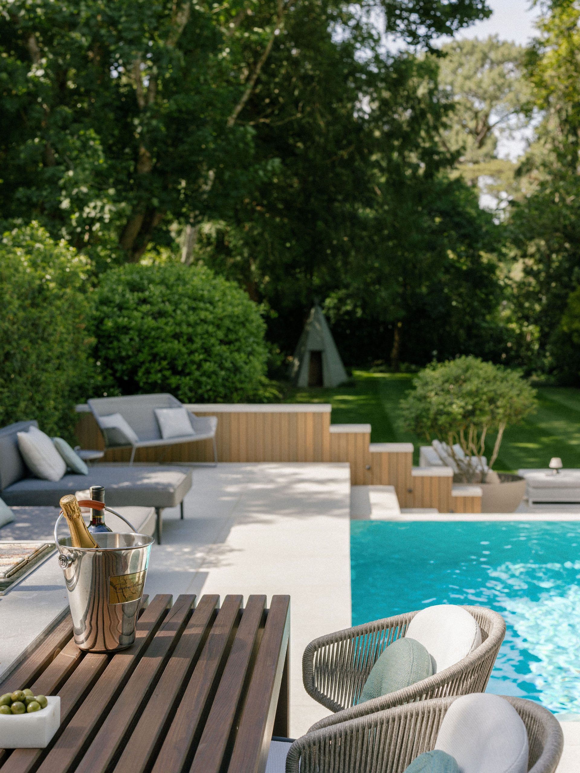 A modern outdoor pool area with loungers, a wooden table, a champagne bucket with bottles, and a bowl of olives. Lush greenery and a small tepee structure are visible in the background.