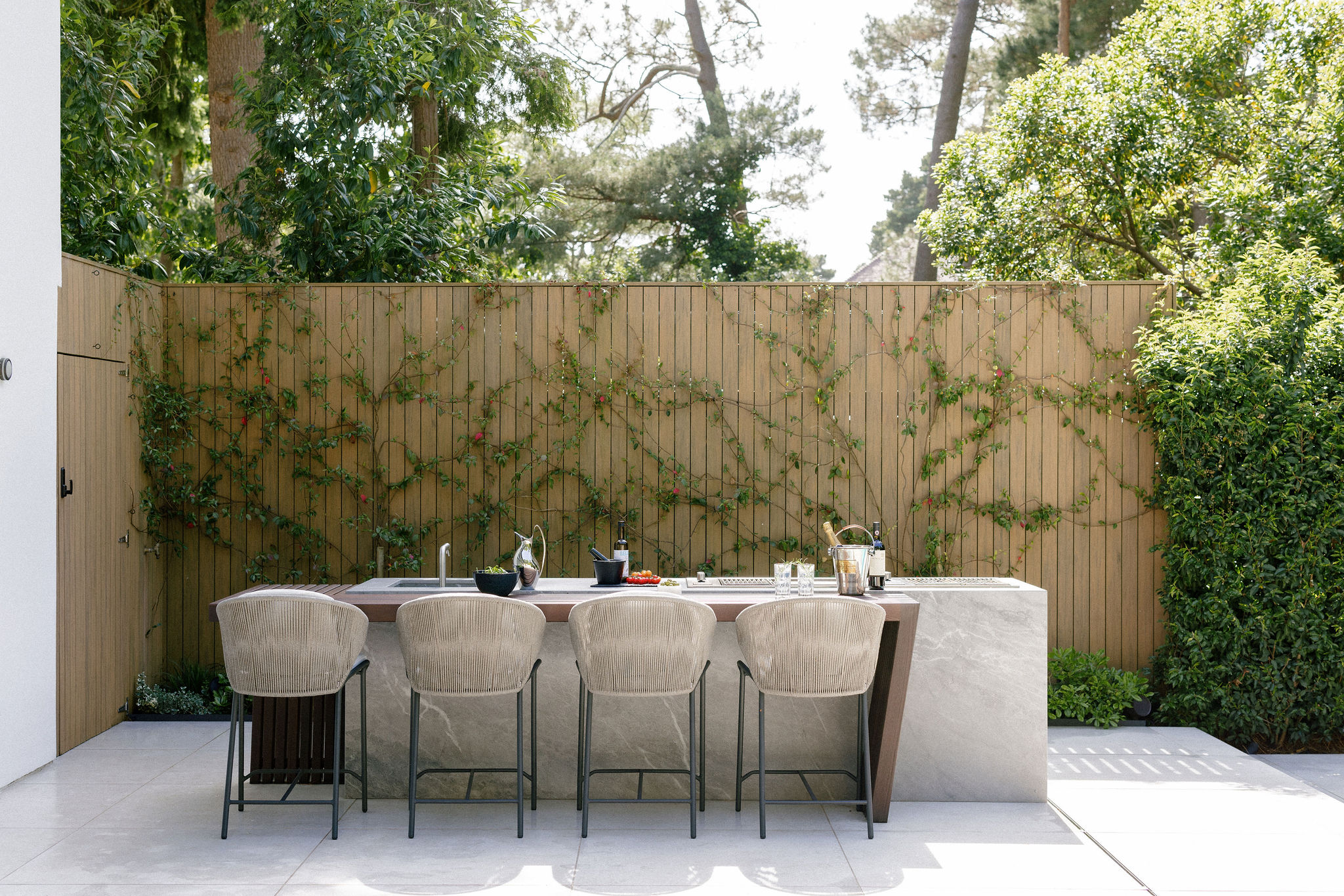 A modern outdoor dining area with a marble-topped table, four woven chairs, and various crockery, set against a wooden fence covered with climbing plants and surrounded by greenery.