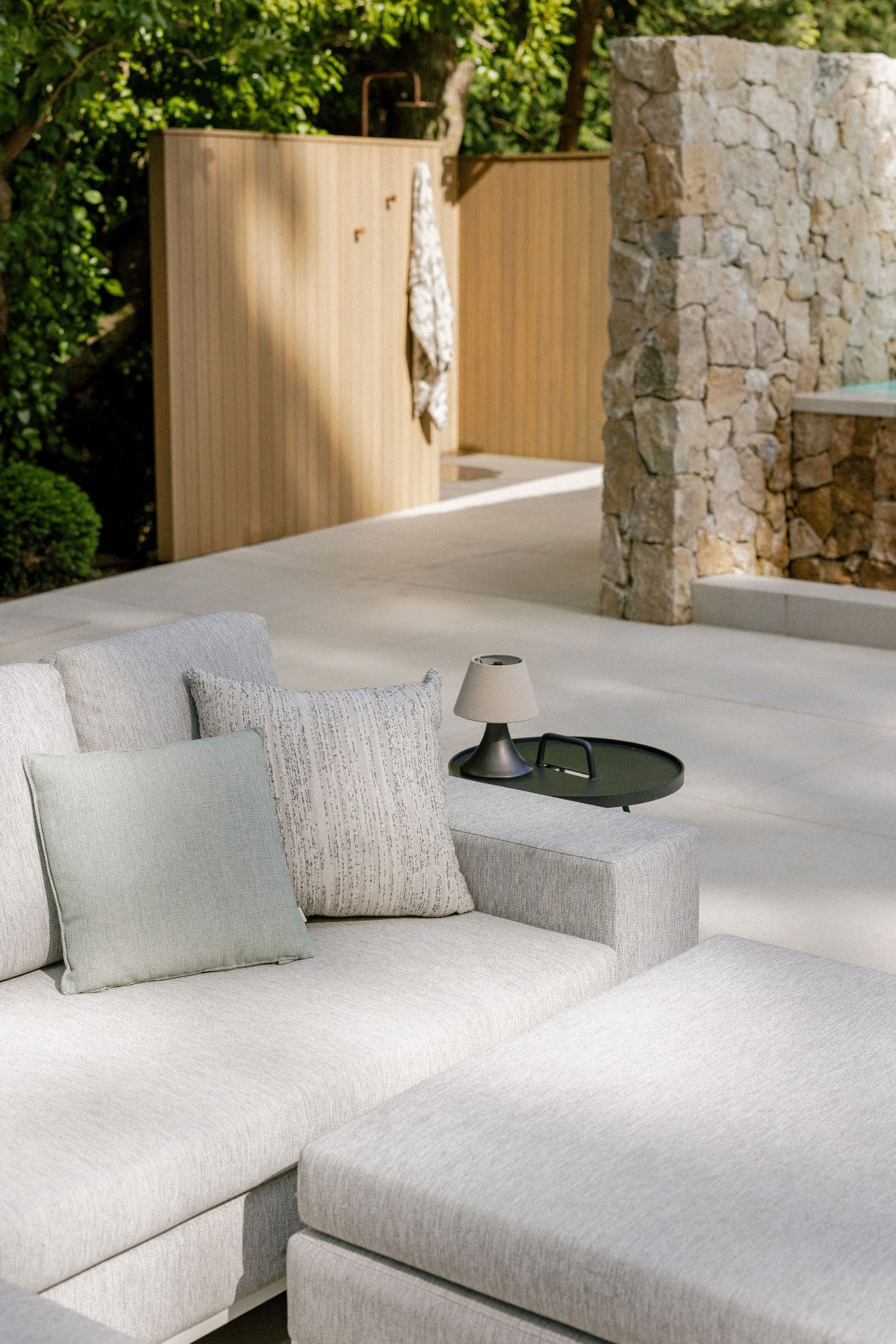 A light grey outdoor sectional sofa with two cushions sits on a patio. A small table with a lamp and tray is nearby. A stone wall and wooden privacy fence are in the background, with a towel hanging on the fence.