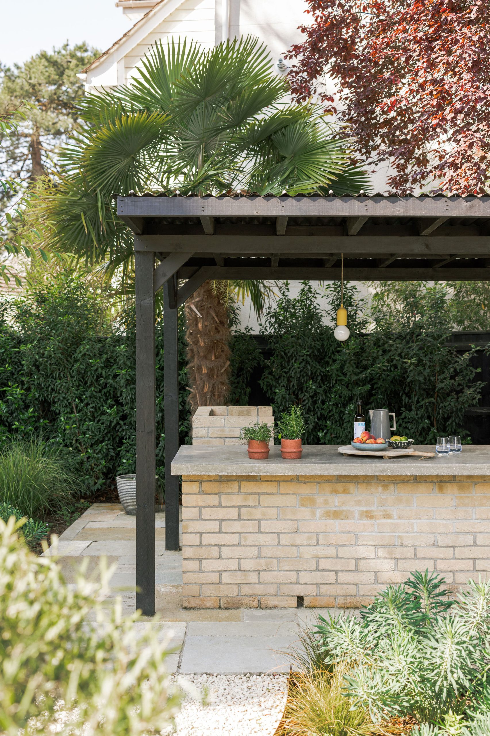 A covered outdoor kitchen with a brick counter holds a few potted plants, a tray with glasses, and a carafe. Lush green plants and a palm tree are visible in the background.
