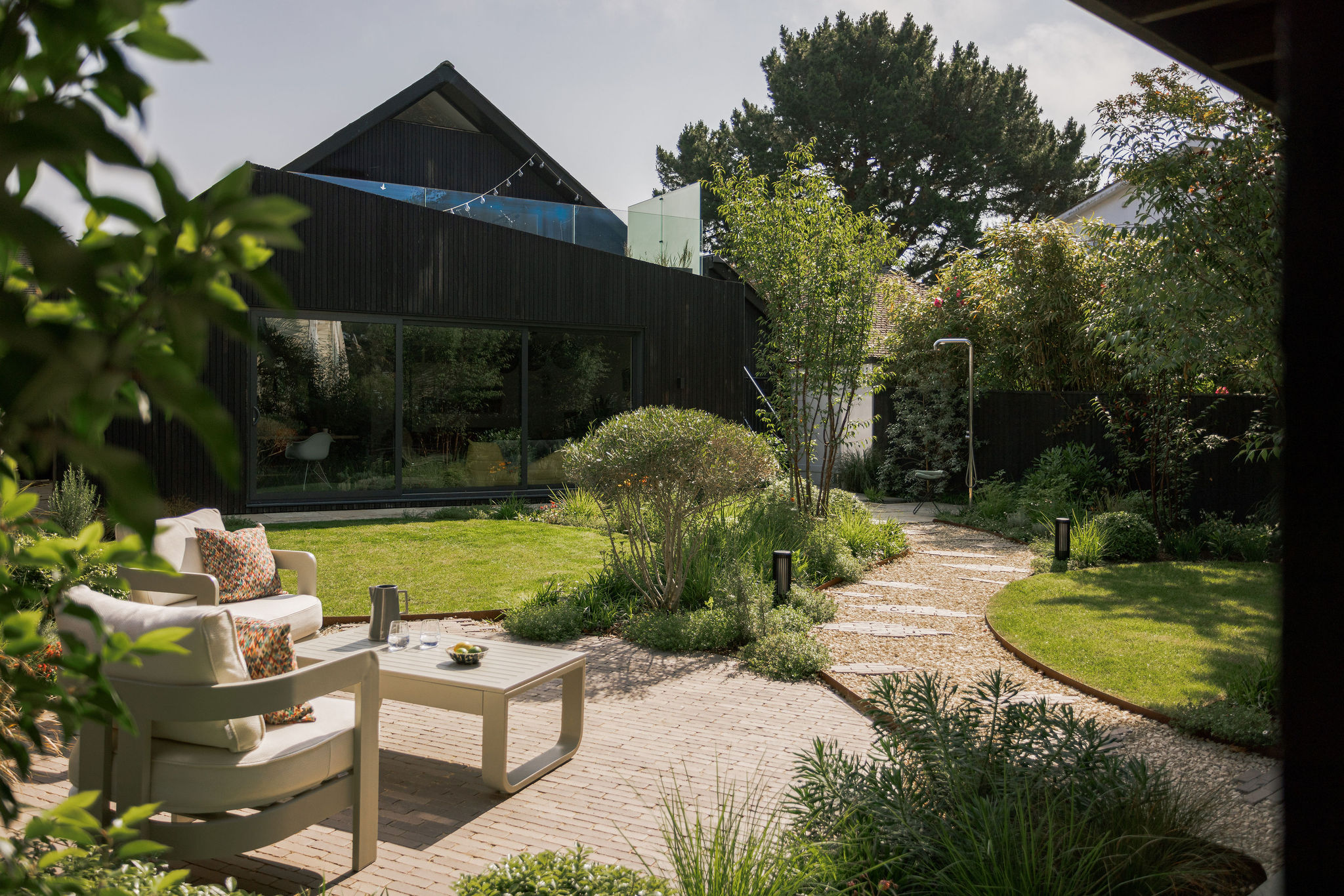 A modern house with dark cladding sits behind a landscaped back garden featuring a paved patio with two cushioned chairs and a table, curving stone path, green lawn, and various plants and trees.
