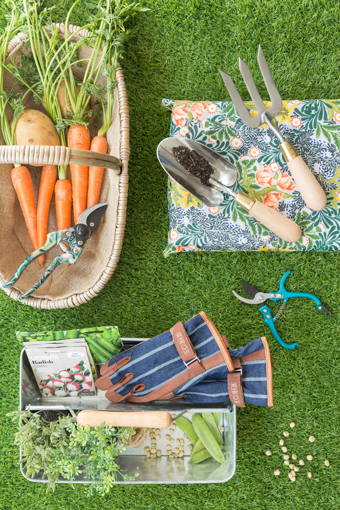 A basket of carrots and potatoes, gardening gloves, secateurs, a hand trowel with soil, a floral kneeling mat, seed packets, peas, and a tray with herbs and garden tools are arranged on green grass.