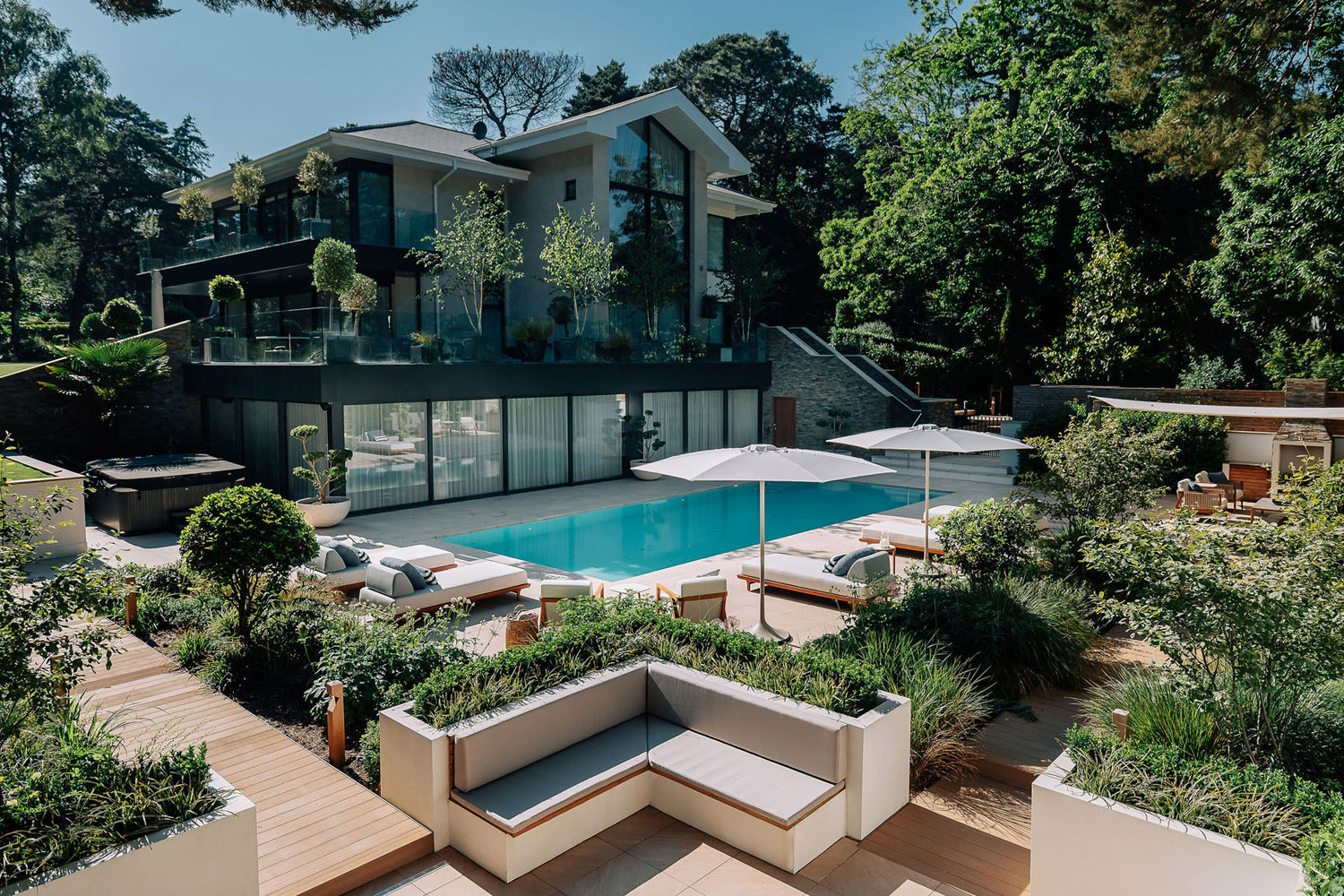 A modern two-storey house with large glass windows overlooks a spacious outdoor area featuring a swimming pool, loungers with umbrellas, landscaped greenery, and built-in seating surrounded by trees.
