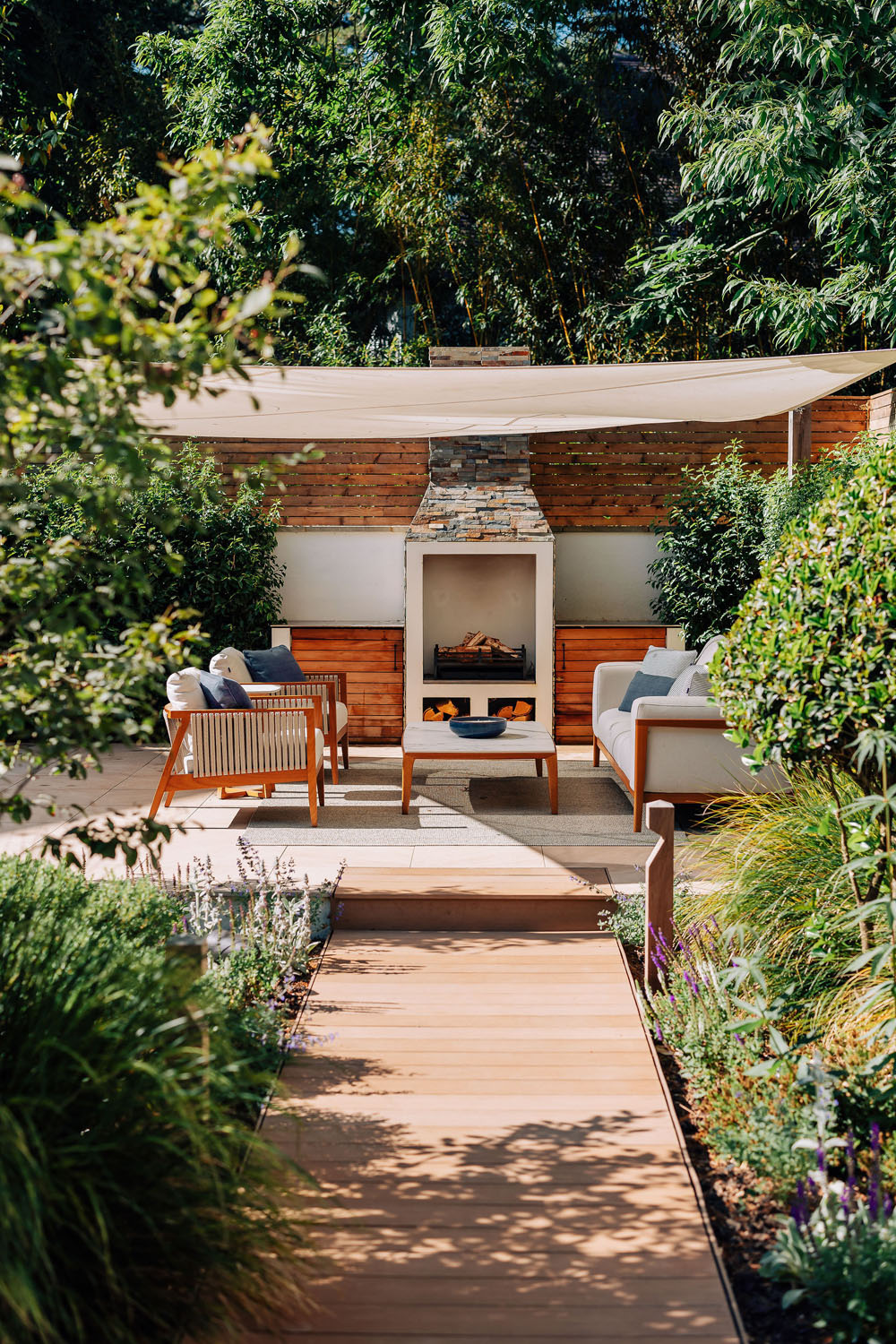 A landscaped garden with a wooden pathway leading to a seating area featuring outdoor sofas, chairs, a coffee table, and a stone fireplace under a white canopy, surrounded by greenery and trees.