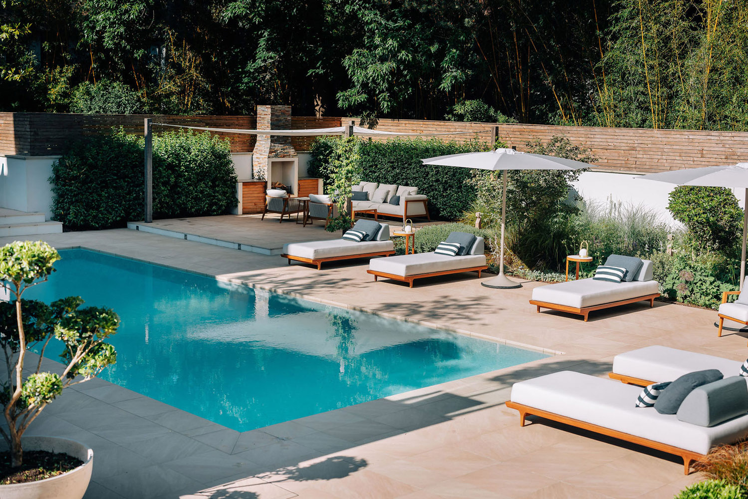 A modern outdoor pool area features several white cushioned sun loungers with parasols, a barbecue grill station, patio tables, and lush greenery surrounding the space. The pool water is clear and calm.