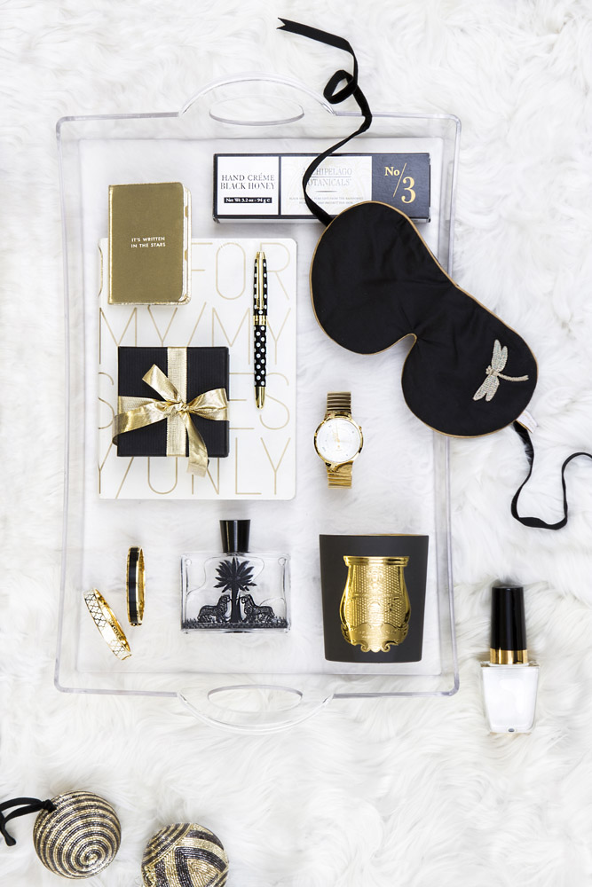 A clear tray on white fur holds a black sleep mask, gold watch, pens, bracelets, a gold candle, perfume, a black gift box with gold ribbon, a notebook, a small book, nail varnish, and striped baubles.