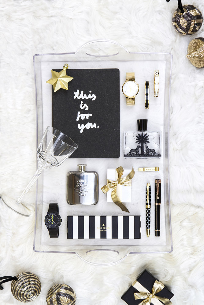 A clear tray on a white fur surface holds a notebook labelled this is for you, a glass, wristwatches, a flask, pens, wrapped gifts with ribbons, a striped box, and decorative ornaments nearby.