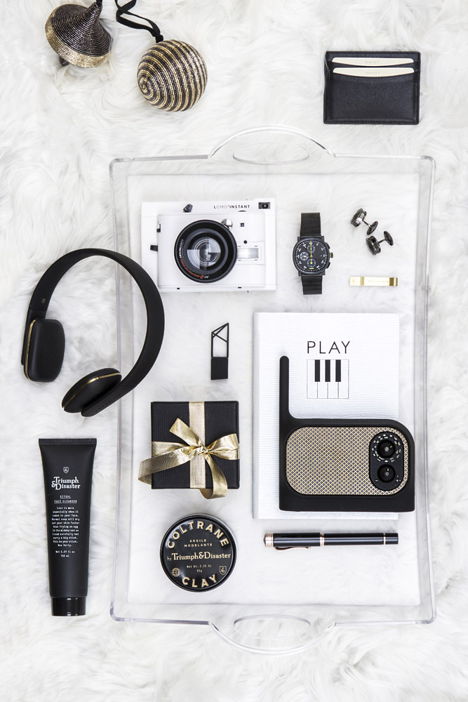 A clear tray holds a camera, black watch, cufflinks, headphones, black tube, wallet, lip balm, notebook, pen, wrapped box, speaker, and small tins, all arranged on a white surface.