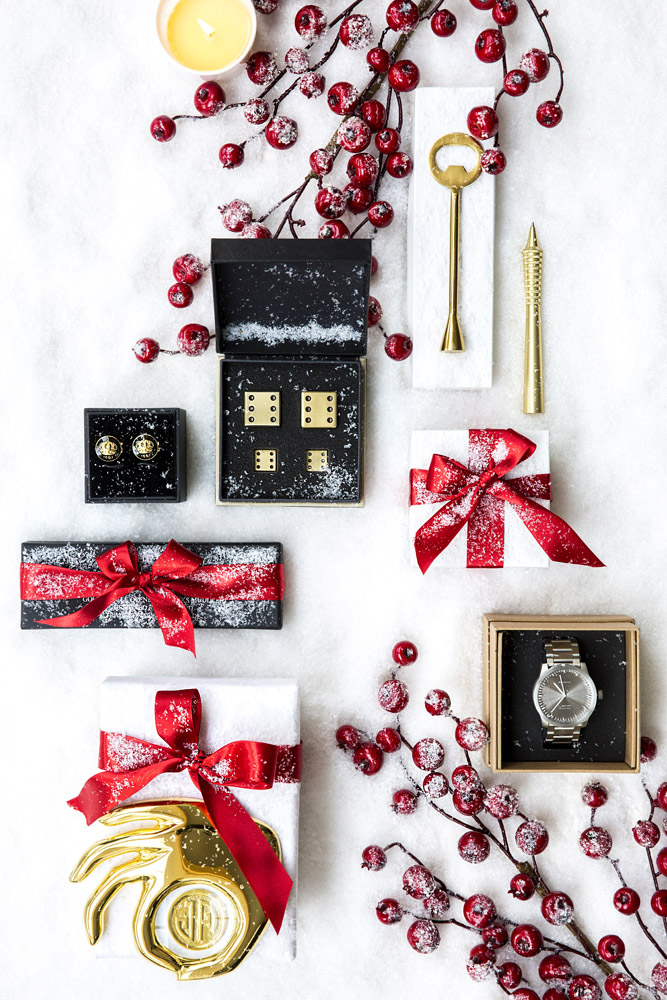 A flat lay of holiday gifts on white fabric with red berry decorations. Items include a watch, gold pen, key, cufflinks, gold dice, a gold bauble, and boxes wrapped with red ribbons, dusted with artificial snow.