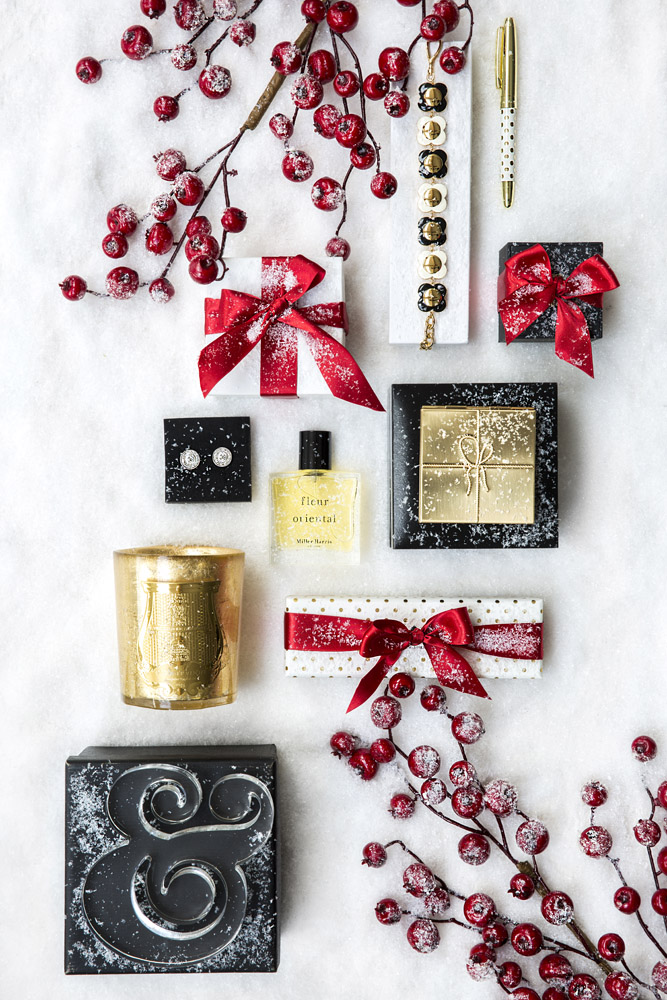 A flat lay of holiday presents on a white surface includes gold and black boxes, red ribbon bows, a perfume bottle, a candle, a pen, jingle bells, and decorative red berries dusted with snow.