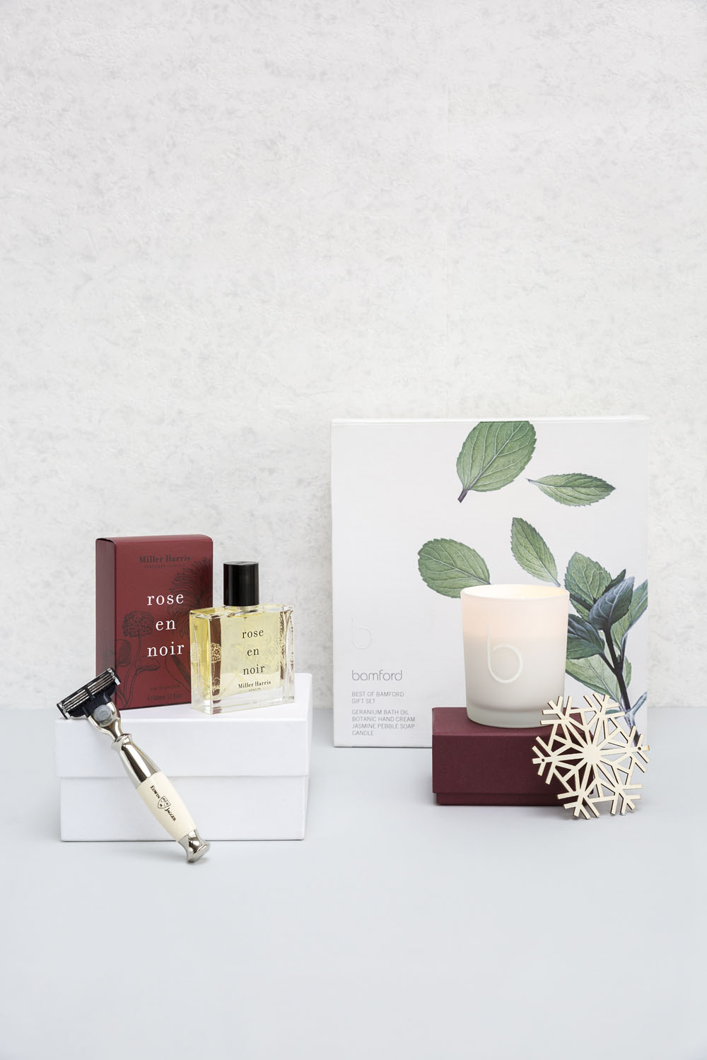 A set of luxury items including a razor, a red box labelled rose en noir, a matching perfume bottle, a white candle in a glass holder, a white box with leaf illustrations, and a decorative white ornament.