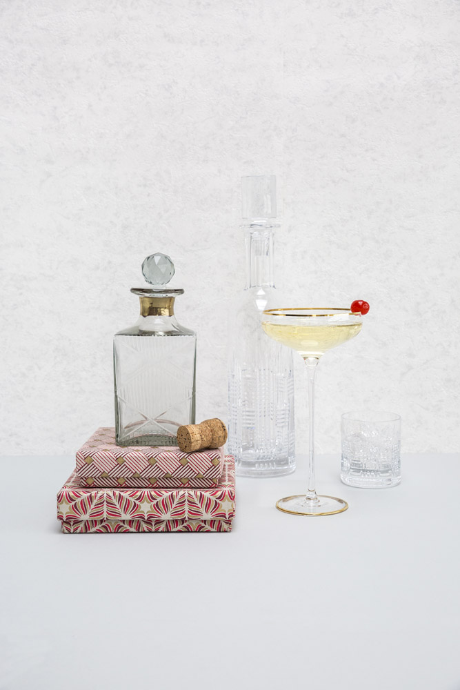 A glass decanter and stopper sit on two patterned books next to a cork, a tall glass bottle, a glass tumbler, and a cocktail glass with a gold rim and a cherry garnish, all on a white surface with a light background.